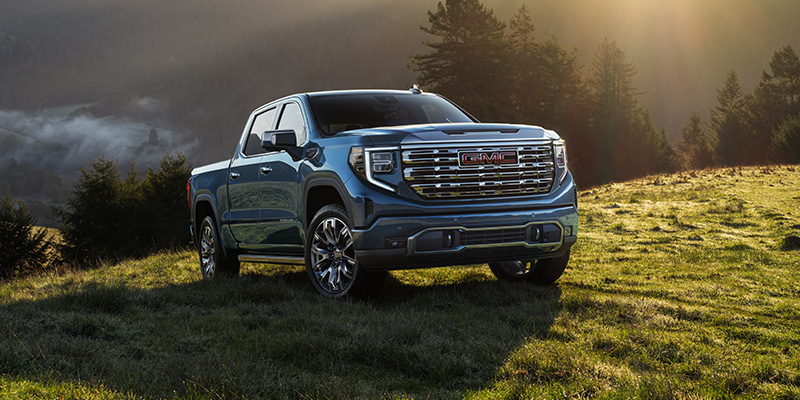 GMC pickup truck parked on a grassy hillside at sunrise, with mountains and trees in the background, showcasing rugged capability and premium design.