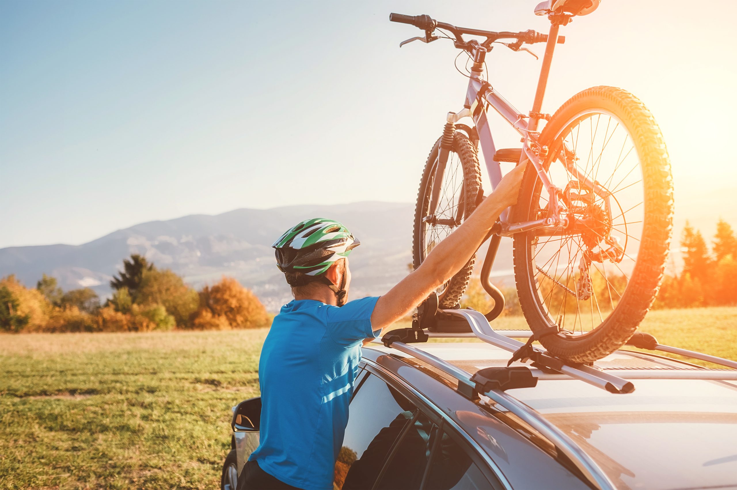 A man in a blue shirt and helmet secures a bicycle to a car roof rack at sunset, with mountains and autumn trees in the background, conveying adventure and freedom.