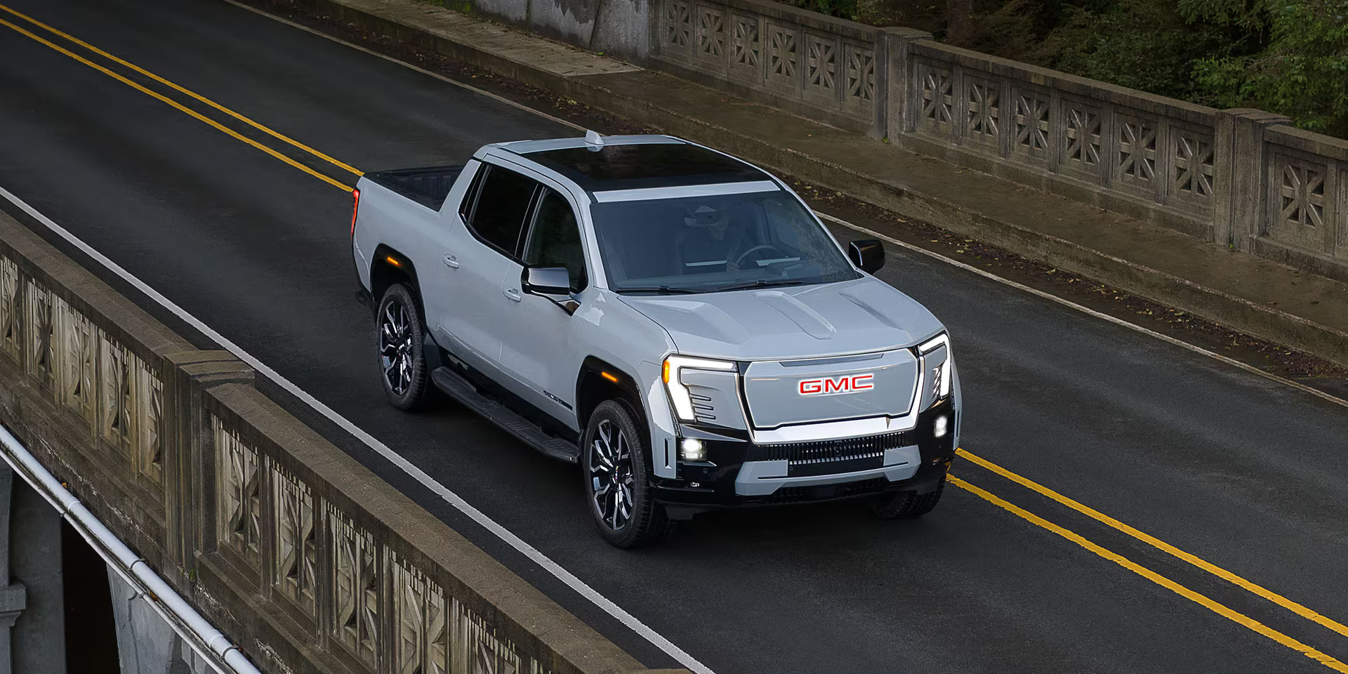 A silver GMC Sierra EV pickup truck driving on a two-lane road beside a stone barrier.