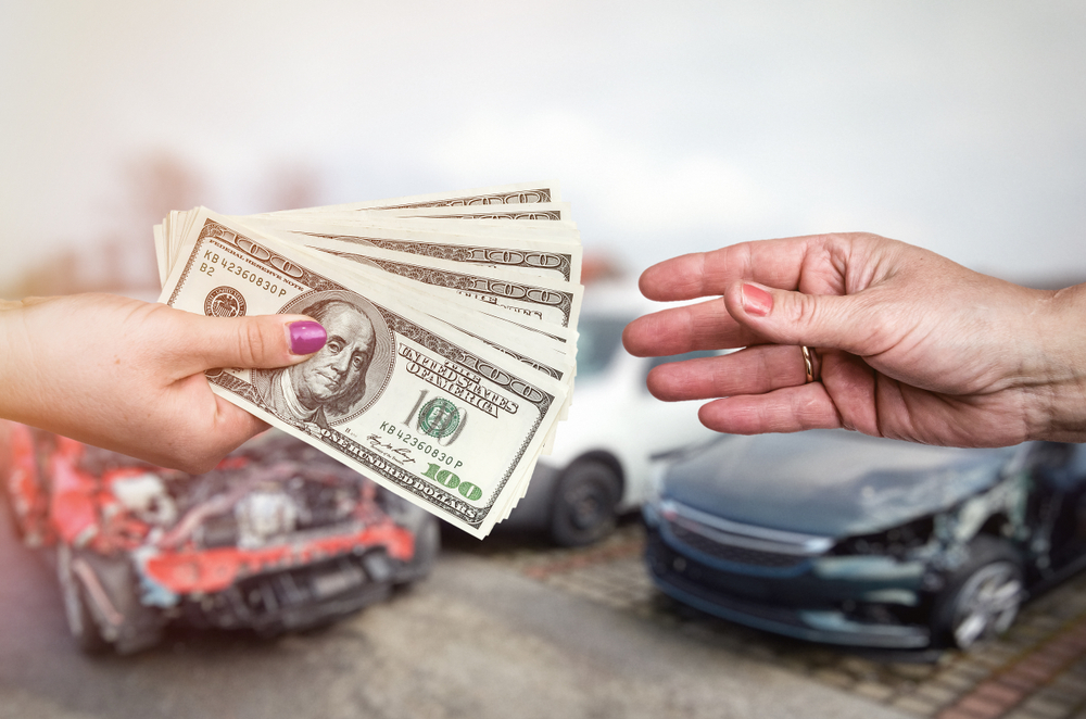 A hand with cash offers money to another hand, with damaged cars in the background.