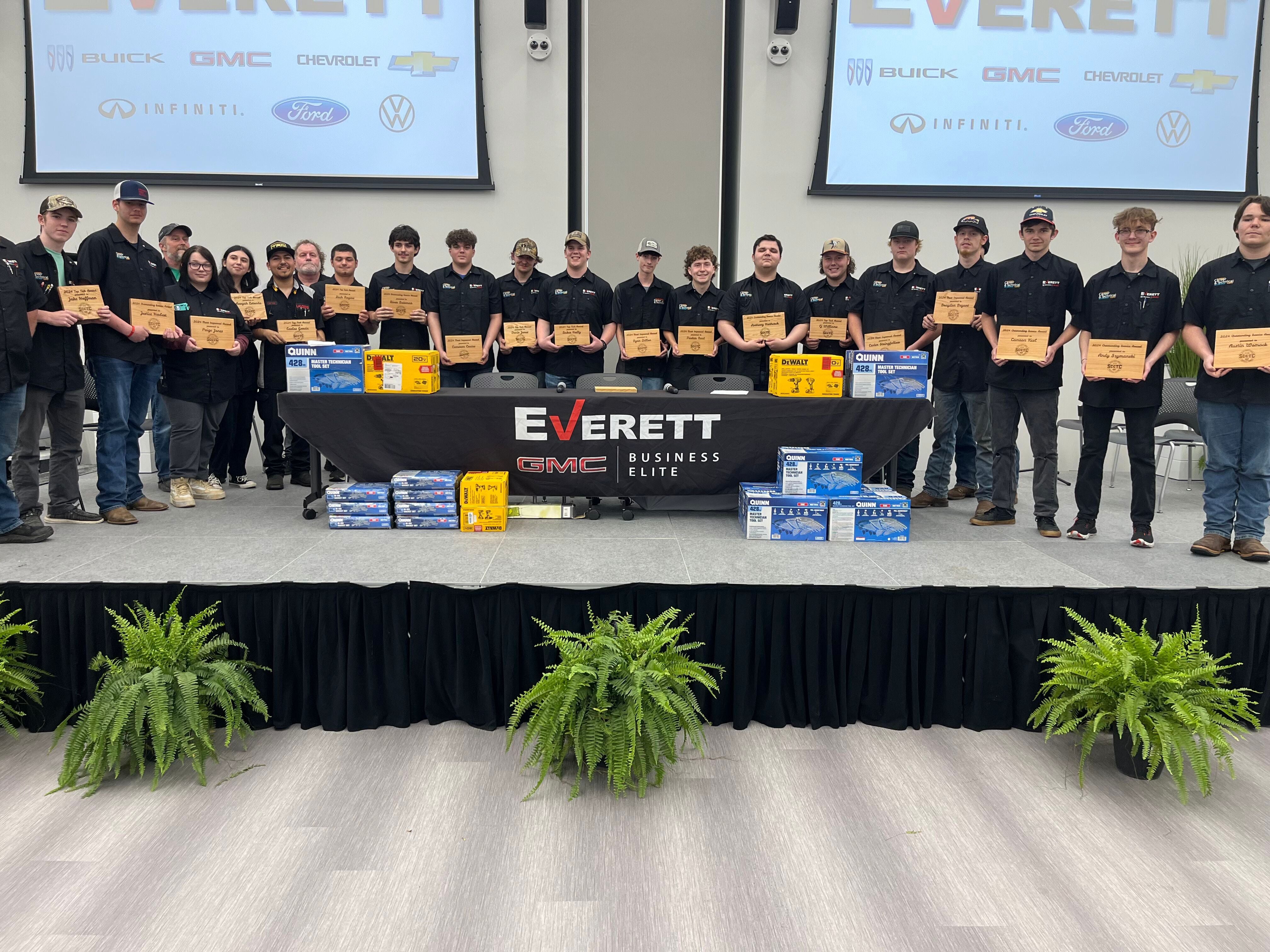 A baseball team in white and red uniforms poses for a group photo on a field, holding a banner that displays the name Everett along with the logos of Buick and GMC. The background shows trees silhouetted against a sunset.