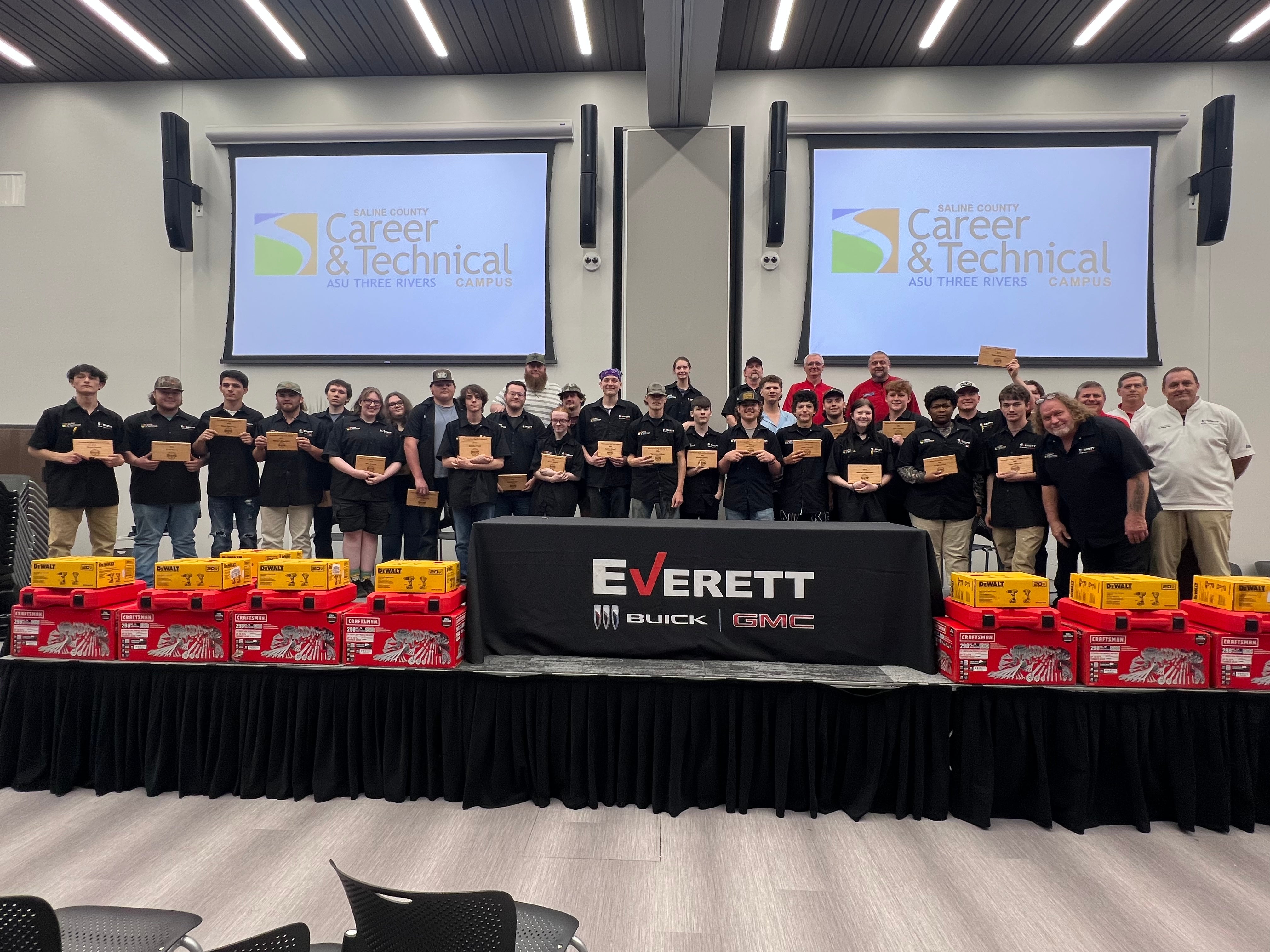 A group of young athletes in baseball uniforms posing for a photo on a field at night, holding a sign that reads Everett.