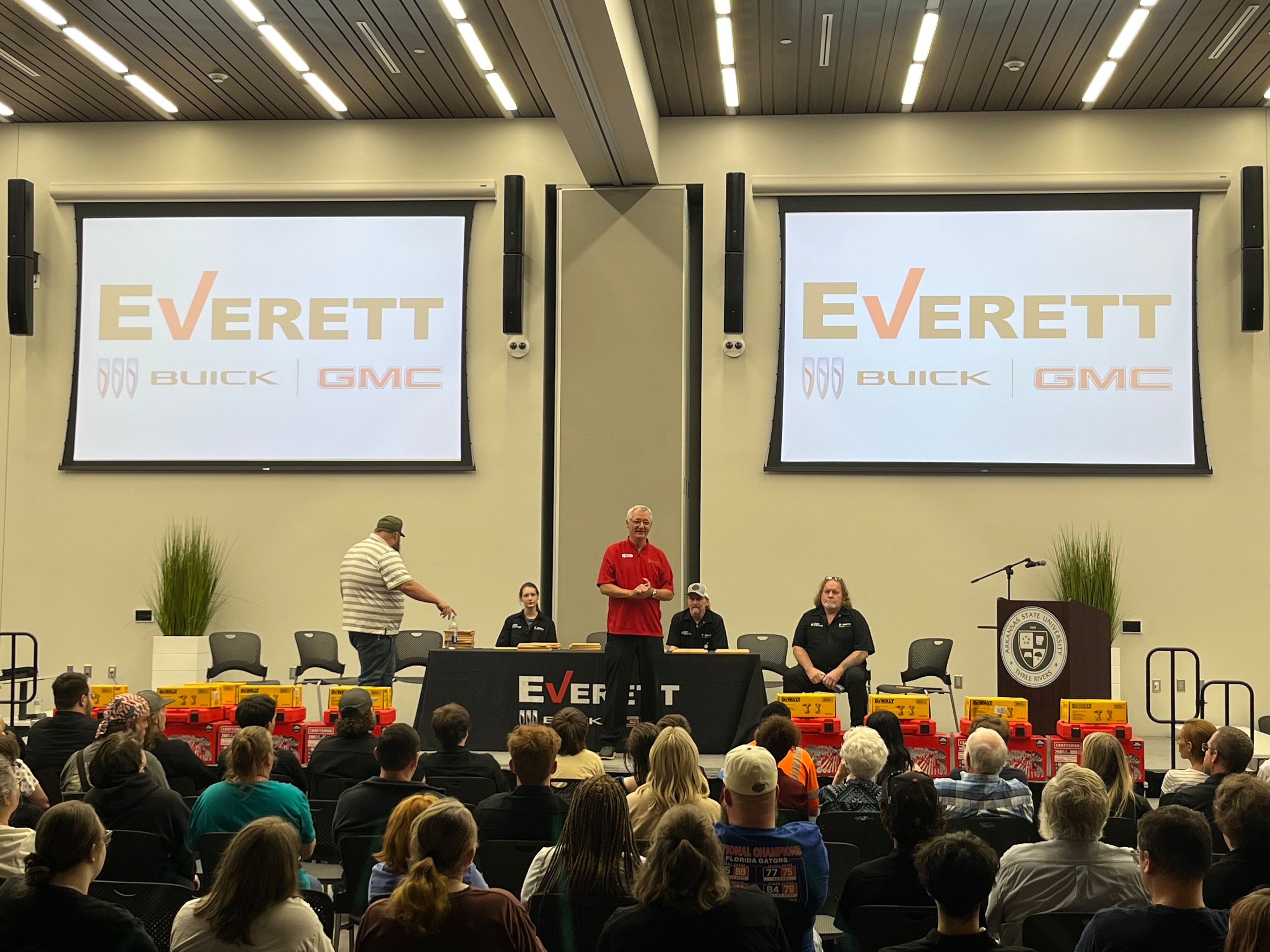 A group of young athletes in baseball uniforms posing for a photo on a field at night, holding a sign that reads Everett.
