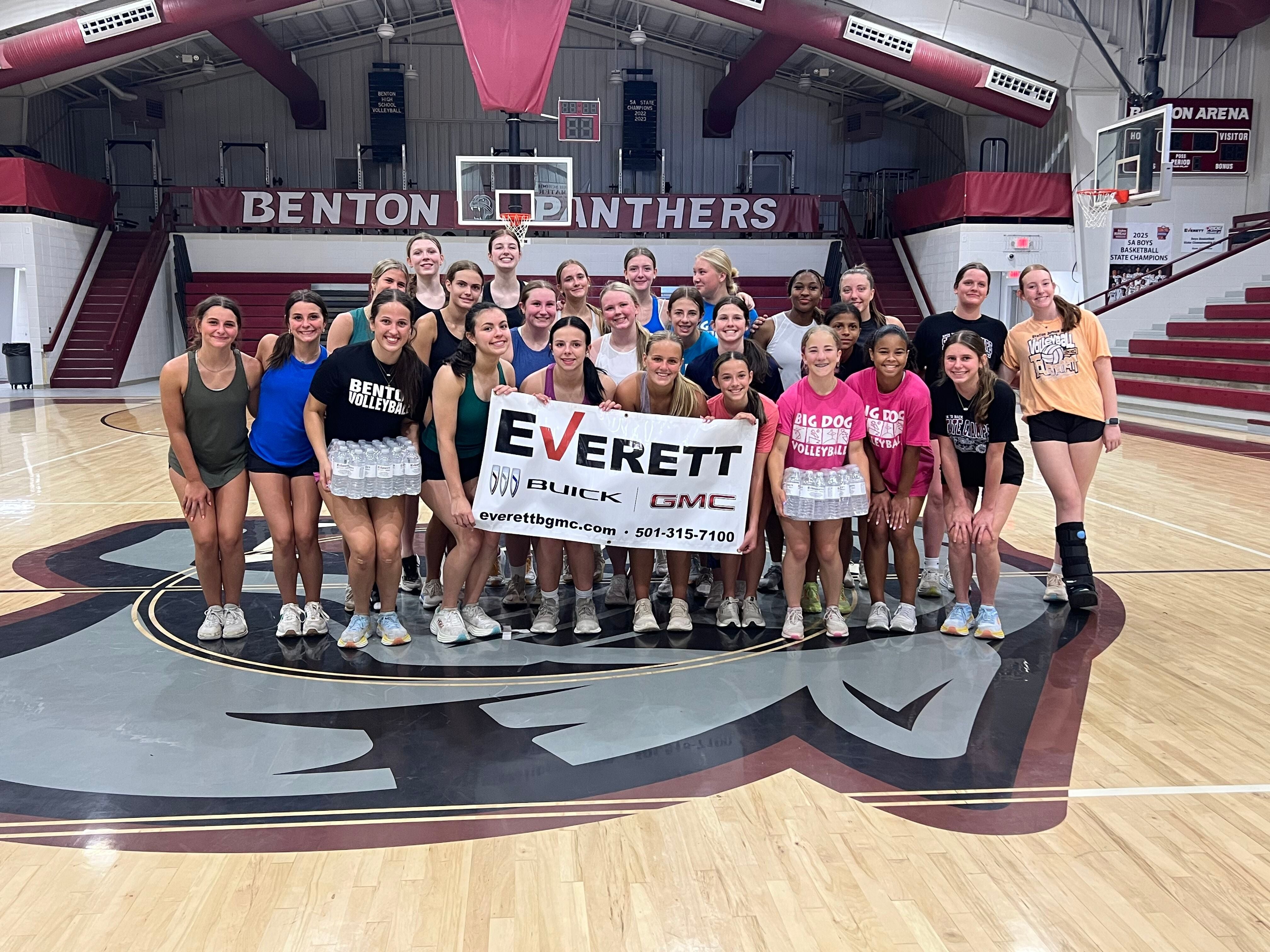 A group of young athletes in baseball uniforms posing for a photo on a field at night, holding a sign that reads Everett.