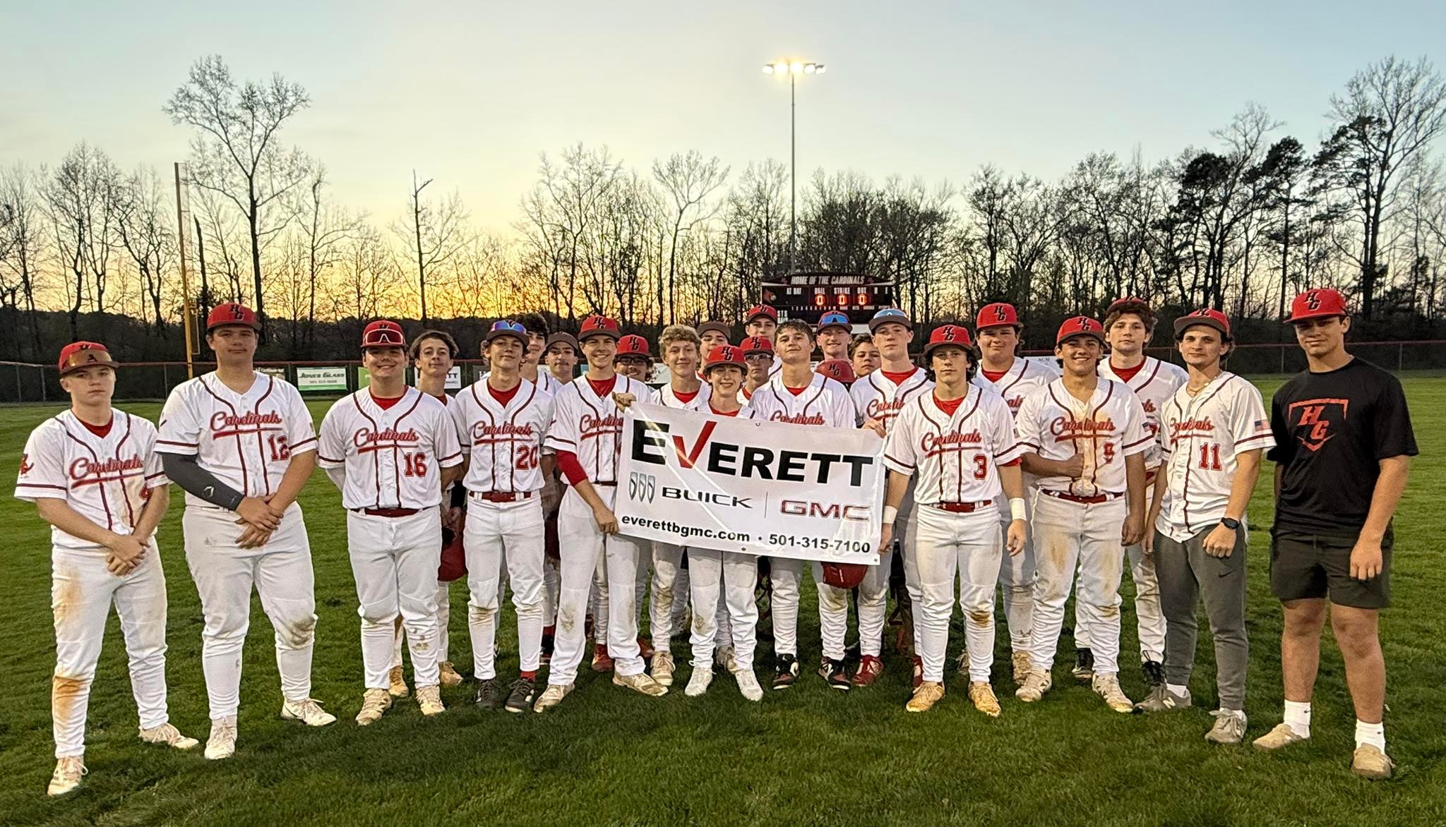 A baseball team in white and red uniforms poses for a group photo on a field, holding a banner that displays the name Everett along with the logos of Buick and GMC. The background shows trees silhouetted against a sunset.
