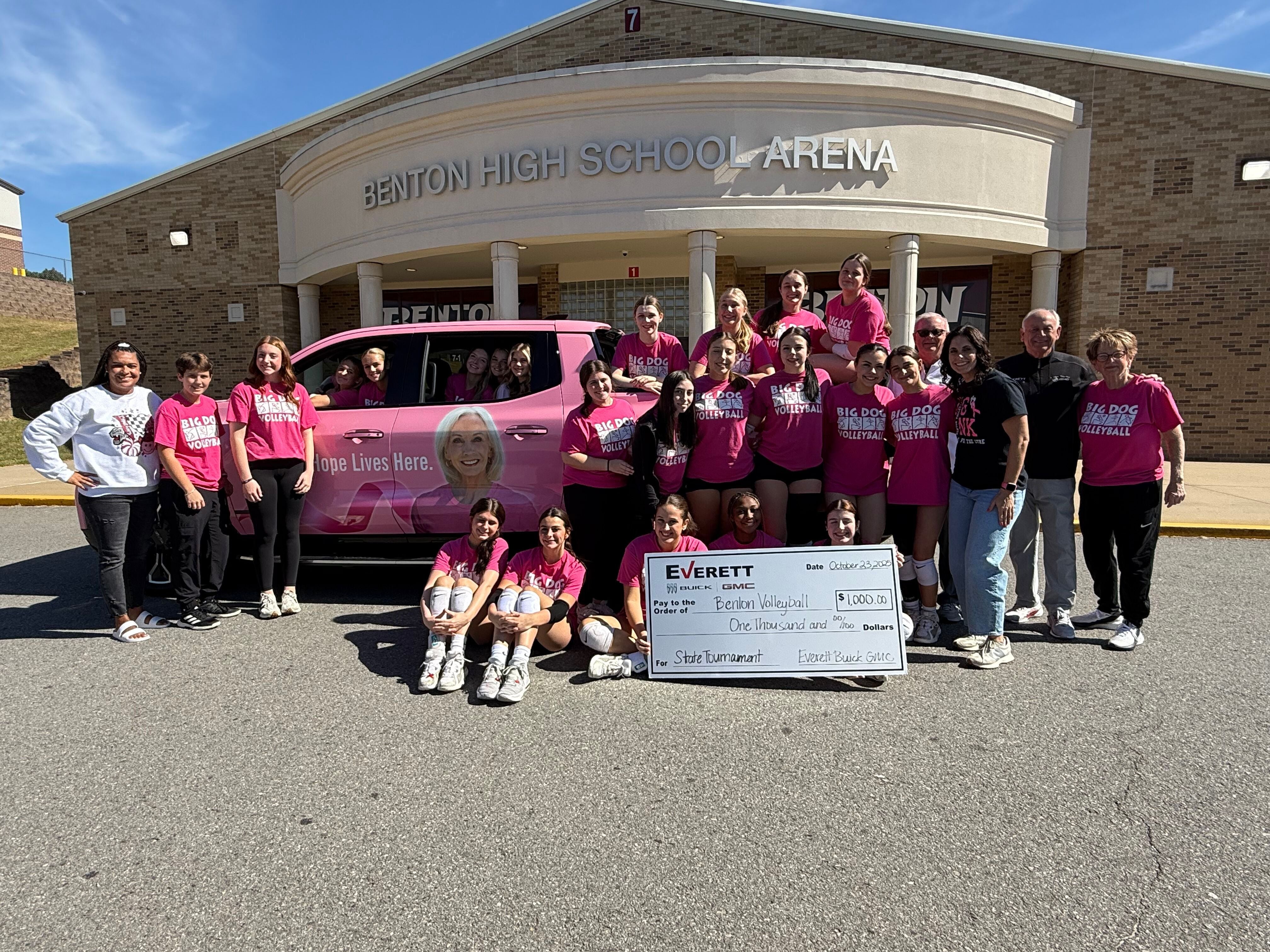 A group of people, including students and adults, poses in front of a pink vehicle and a large check at Benton High School Arena.