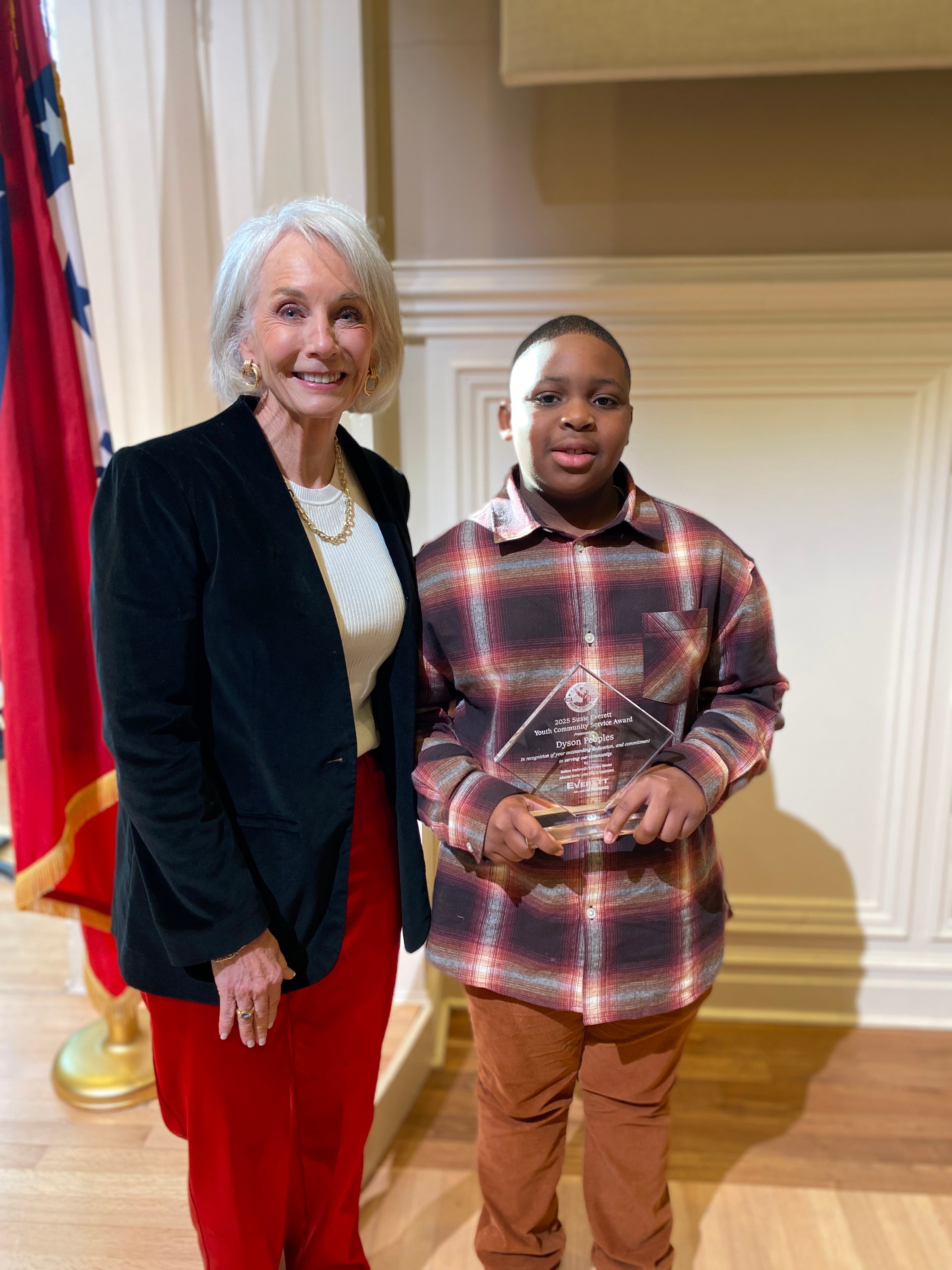 A woman in a blazer and jewelry stands next to a young boy holding an award. They are posed in front of a flag and an indoor setting.