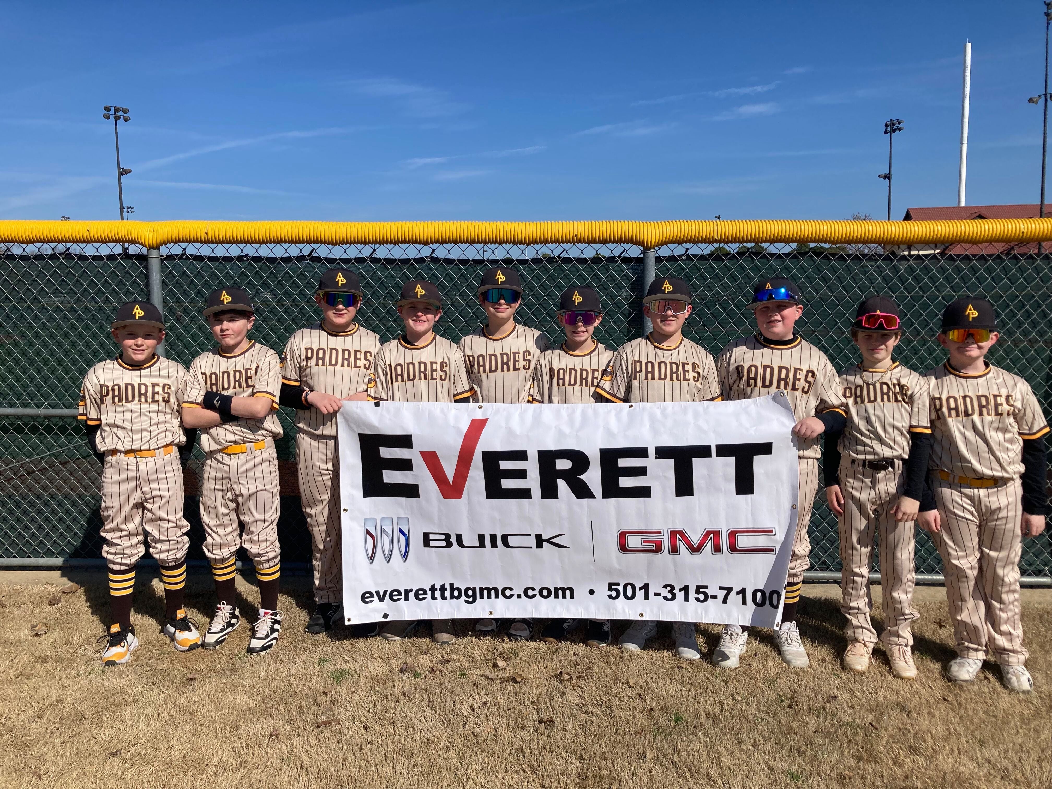 A group of young baseball players wearing beige jerseys with PADRES written on them, standing in front of a fence and holding a large banner that says Everett Buick GM along with contact information.