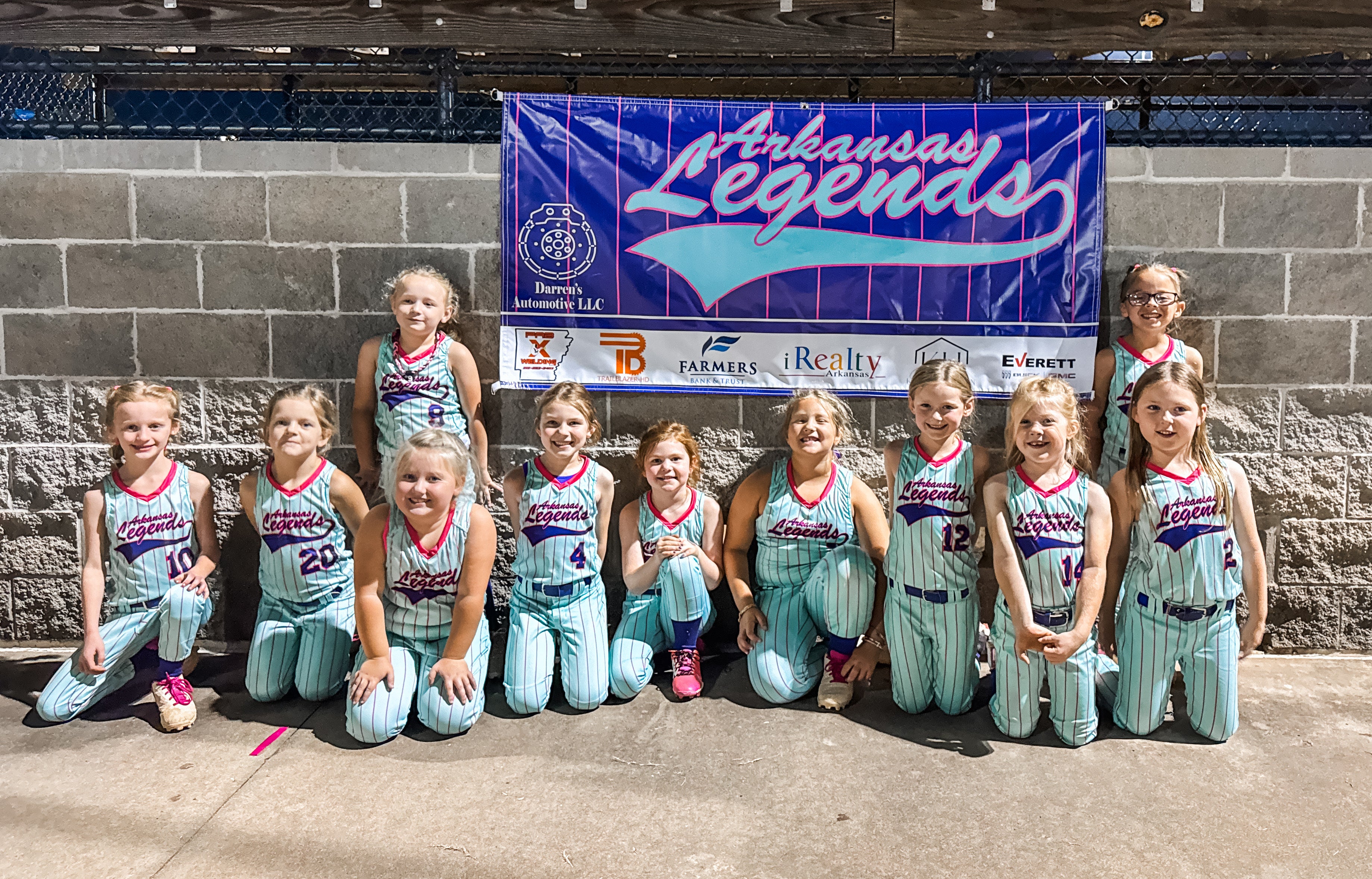 Youth softball team posing together in uniform under a team banner.